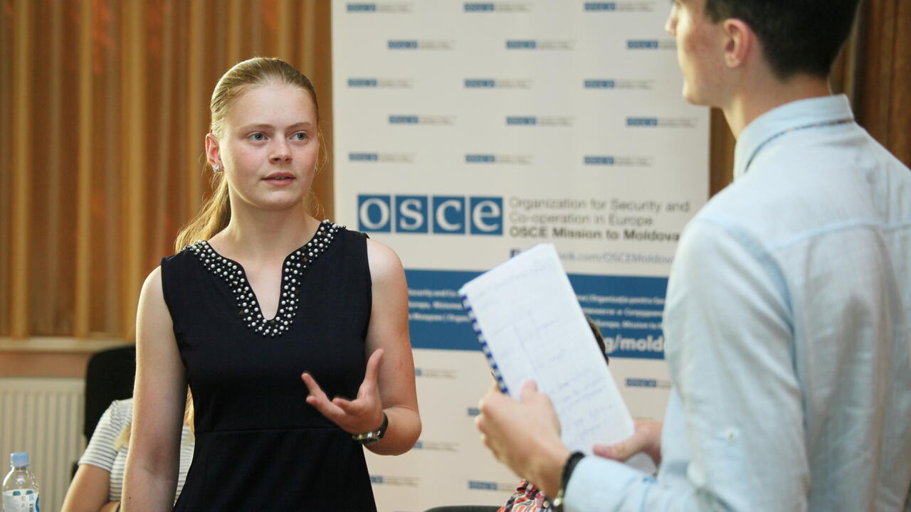 Two people engaged in discussion at an event, with a logo banner in the background.